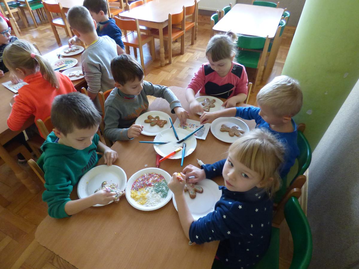 Przedszkolaki siedzą przy stoliku, malują lukrem i dekorują pisakami cukrowymi i różnego rodzaju posypkami pierniczki.