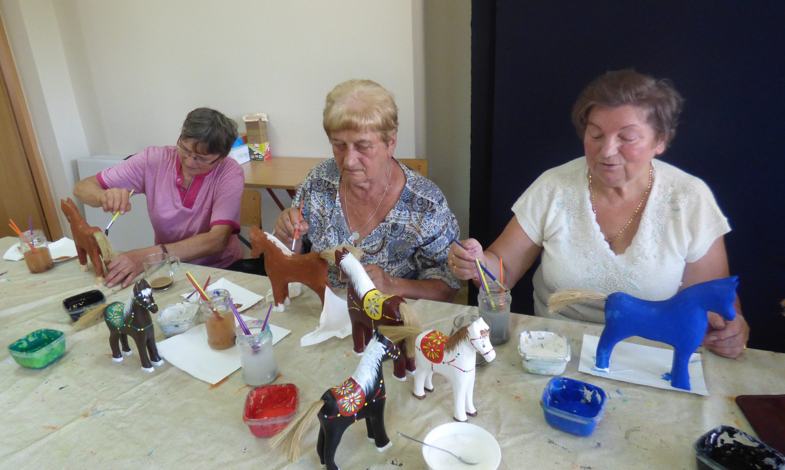 Zdjęcie przedstawia seniorów malujących drewniane koniki.