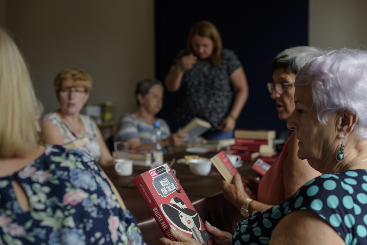 Zdjęcie przedstawia pięć siedzących i jedną stojącą kobietę przy stolena świetlicy GOK. Dwie z nich trzymają w ręku książkę autorki Louise Walters „Walizka Pani Sincilar”. Widoczne na zdjęciu kobiety dyskutują na temat przeczytanej już książki, pt. „Jak zawsze”.