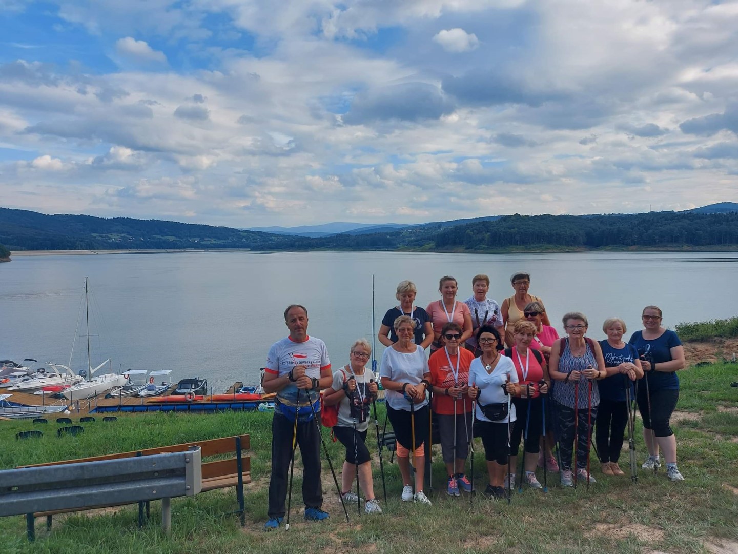 Czternastu uczestników spaceru z kijkami stoi pozując do wspólnego zdjęcia. Za ich plecami widać Jezioro Mucharskie.