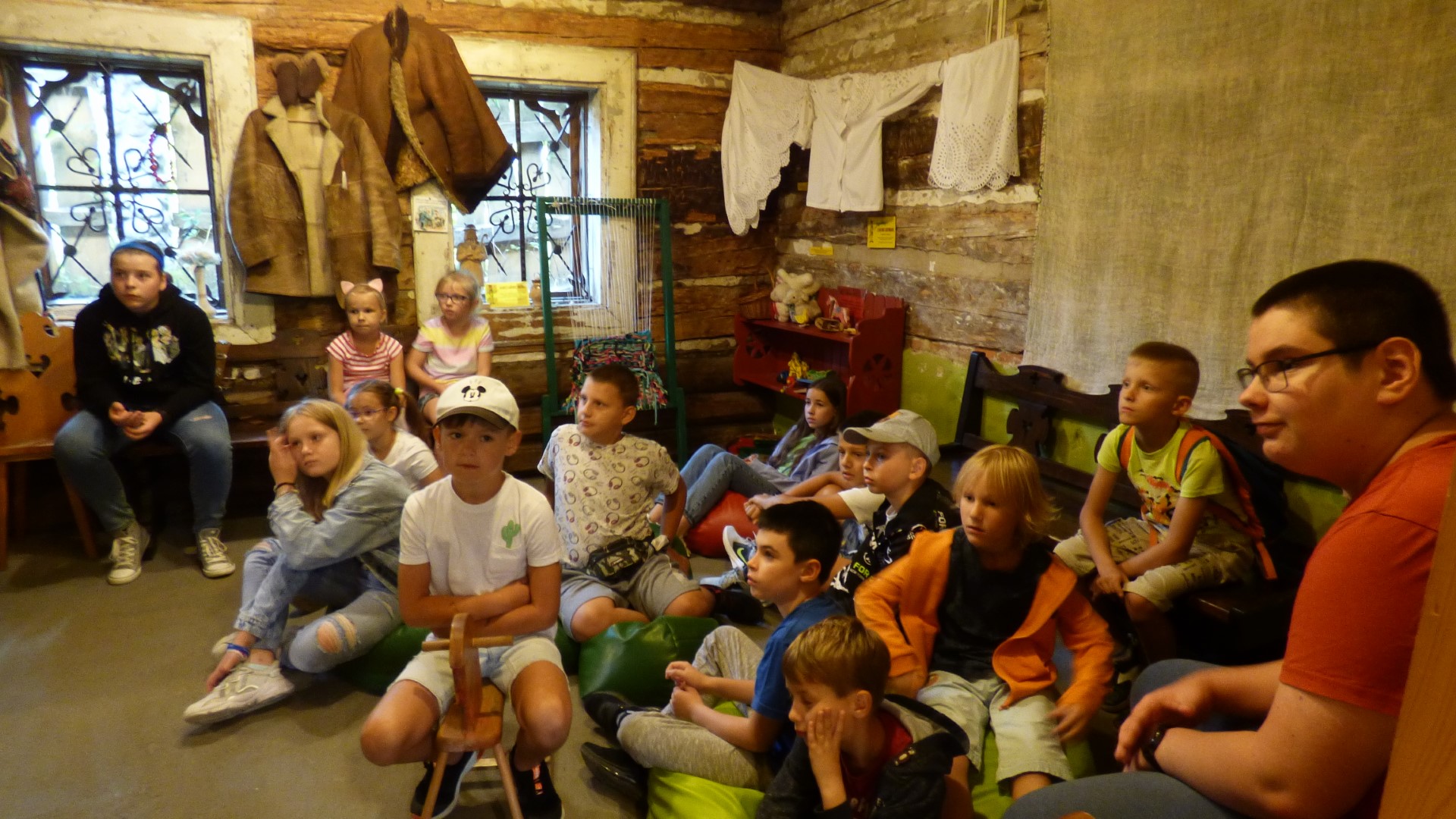 Uczestnicy wycieczki siedzą na podłodze lub ławkach w izbie góralskiej chaty i słuchają opowiadania pani przewodnik.