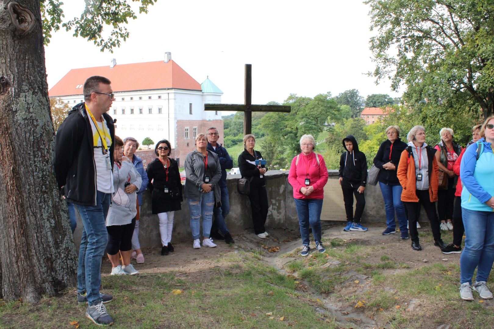 Uczestnicy wycieczki stoją obok Katedry w Sandomierzu i słuchają stojącego z lewej strony przewodnika, w tle widać zamek królewski.
