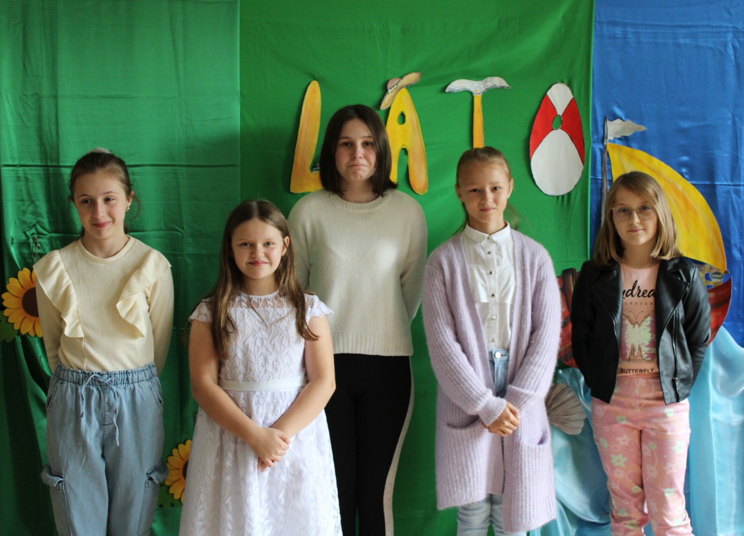 Zdjęcie przedstawia pięć dziewczynek pozujących do zdjęcia grupowego podczas Konkursu Recytatorskiego „Cztery Pory Roku – LATO” organizowanego przez Gminny Ośrodek Kultury w Stryszowie. Dzieci stoją na tle zielono-niebieskiej dekoracji, nad nimi widnieje żółty napis LATO
