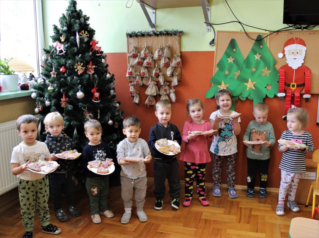 Dziewięcioro przedszkolaków z grupy „Maluszków” stoi na tle choinki i pozuje do wspólnego zdjęcia trzymając w rękach talerze z pomalowanymi pierniczkami.