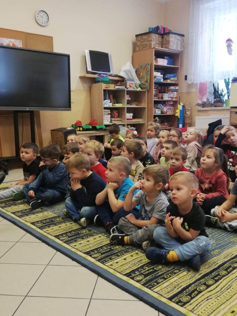 Przedszkolaki siedzą na dywanie zasłuchane w czytanie pana bibliotekarza.