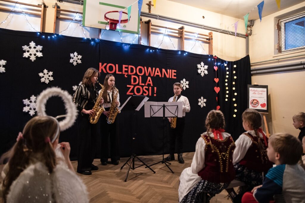 Na zdjęciu znajdują się dwie dziewczynki i jeden chłopiec ze szkoły muzycznej w Kalwarii Zebrzydowskiej, którzy grają na saksofonach kolędę, podczas Kolędowania dla Zosi Pająk, zorganizowanego przez Gminny Ośrodek Kultury w Stryszowie.