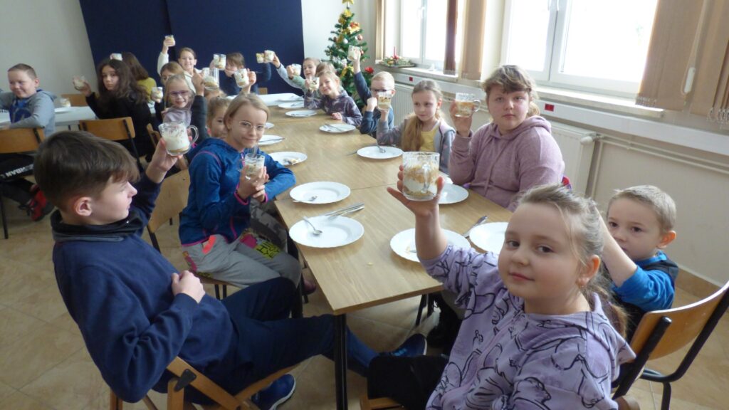 Na zdjęciu znajduje się dziewiętnaście dzieci, które siedzą na krzesełkach przy stolikach i trzymają uniesione w ręce deserki w szklankach, które przyrządzili na Kąciku Kulinarnym zorganizowanym przez Gminny Ośrodek Kultury w Stryszowie podczas Ferii zimowych.