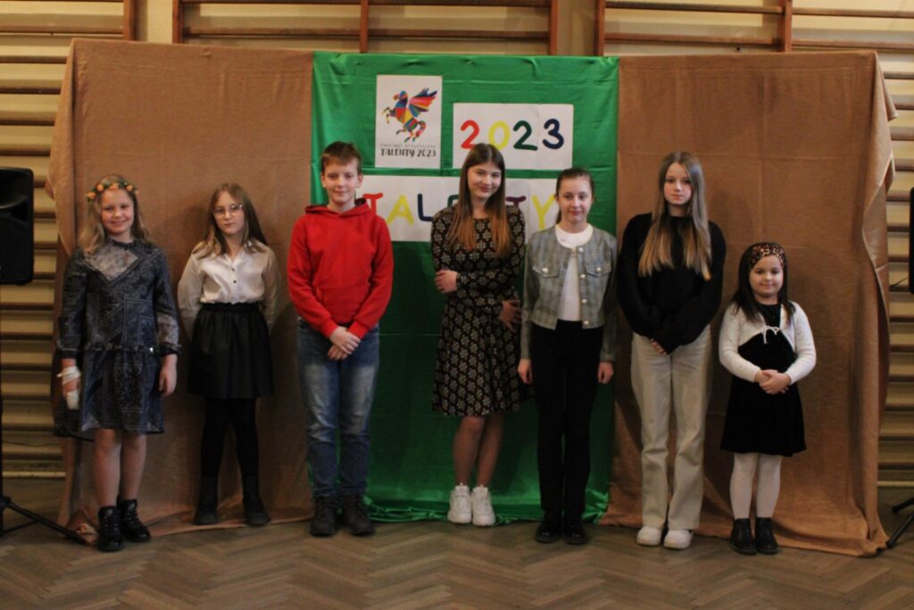 Na zdjęciu znajduje się sześcioro dziewczynek i jeden chłopiec, którzy pozują do zdjęcia na małej Sali gimnastycznej w Stroniu podczas przesłuchań konkursowych etapu gminnego Festiwalu Artystycznego „TALENTY 2023” w kategorii wokal.
