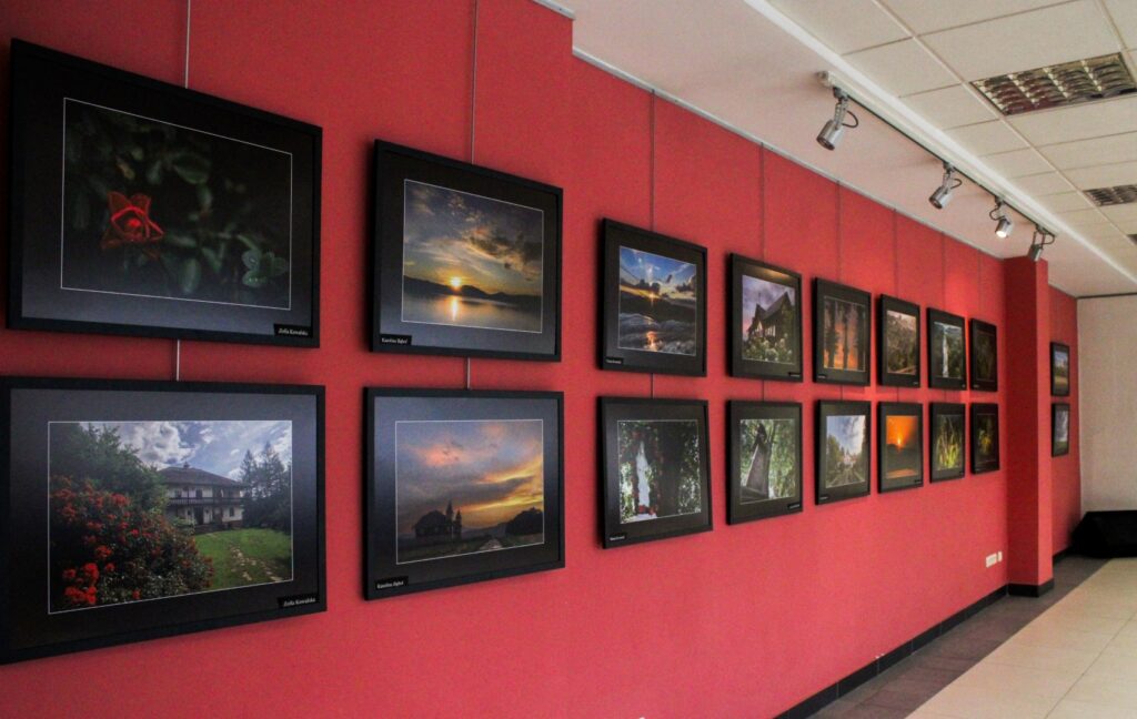 Zdjęcie przedstawia wystawę, gdzie na tle czerwonej ściany w Wadowickiej Bibliotece Publicznej zawieszone są zdjęcia z projektu realizowanego przez Gminny Ośrodek Kultury w Stryszowie - „Stryszów – dotyk piękna”.