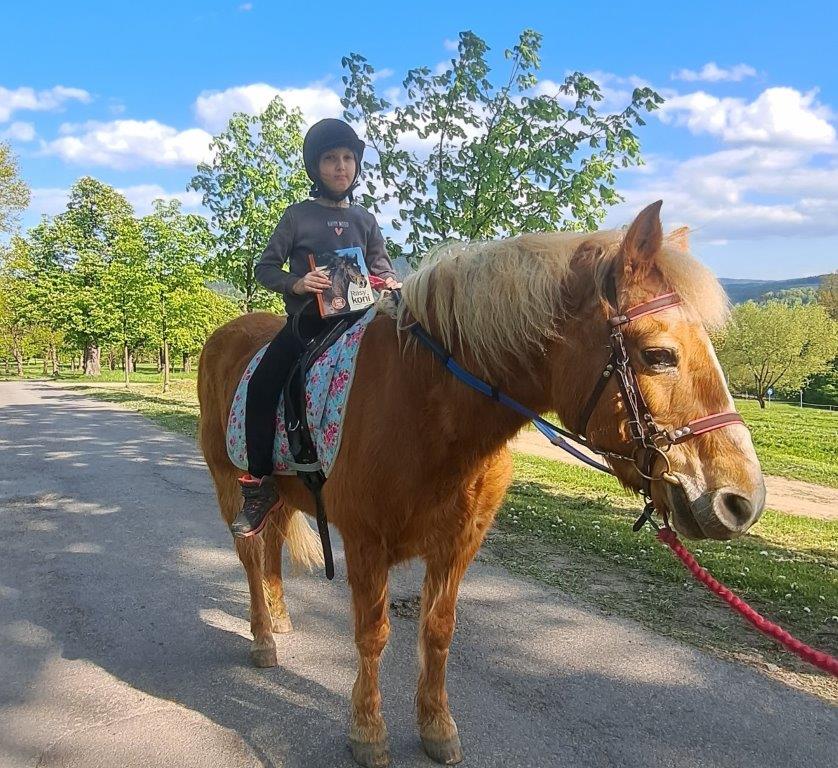Fotografia przedstawia dziewczynkę w kasku jeździeckim siedzącą na koniu i trzymającą w ręku książkę. Praca została wysłana na konkurs fotograficzny organizowany przez biblioteki publiczne gminy Stryszów „Z książką mi do twarzy”.