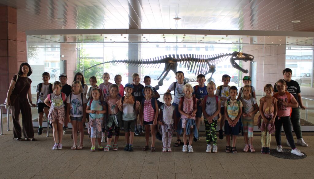 – Dzieci uczestnicy wycieczki wraz z opiekunem stoją przed szkieletem dinozaura i pozują do wspólnego zdjęcia.