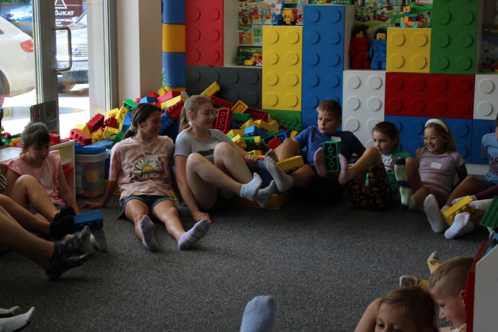 Zdjęcie przedstawia grupę dzieci siedzącą w kole, w Edukacyjnej Sali Zabaw AleKlocki w Myślenicach podczas zabawy polegającej na podawaniu sobie dużych klocków stopami