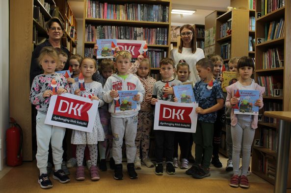Grupa dzieci stoi w bibliotece. Trzymają plakaty z logiem Dyskusyjnego klubu książki