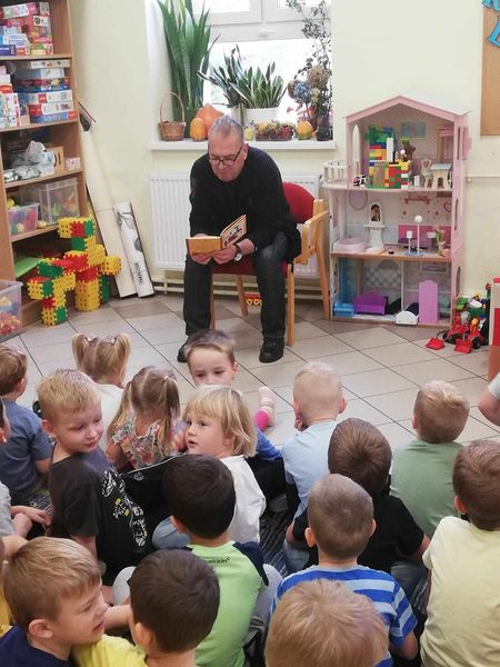 Na krzesełku w przedszkolu siedzi mężczyzna wokół niego małe dzieci. Mężczyzna czyta książkę.