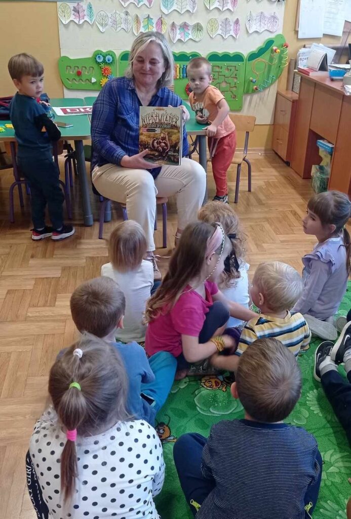 Na krzesełku w przedszkolu siedzi kobieta wokół niej małe dzieci. Kobieta trzyma w rękach książkę.