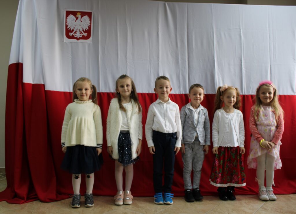 cztery dziewczynki i dwóch chłopców. Pozują oni do zdjęcia grupowego podczas konkursu muzycznego „Piosenka Patriotyczna”z klasy zero. W tle flaga Polski i godło