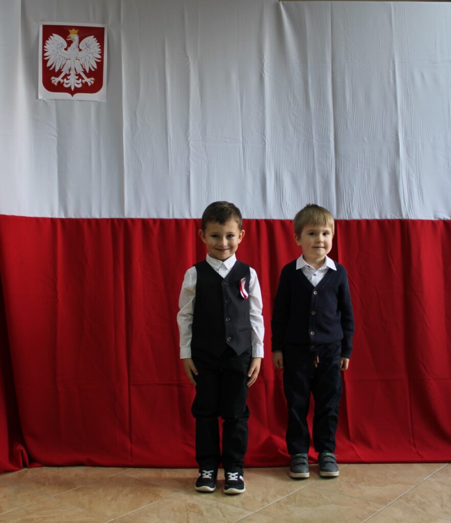 dwóch chłopców. Pozują oni do zdjęcia grupowego podczas konkursu muzycznego „Piosenka Patriotyczna” z kategorii „Maluszki”. W tle flaga Polski i godło