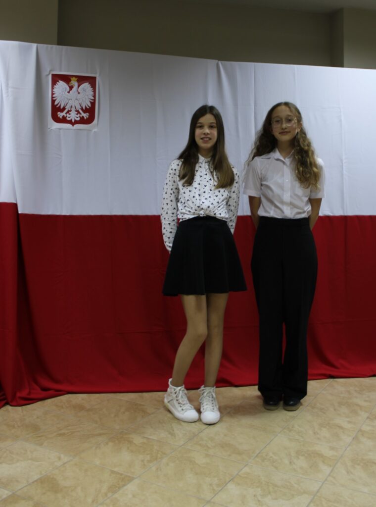 się dwie dziewczynki. Pozują one do zdjęcia grupowego podczas konkursu muzycznego „Piosenka Patriotyczna” z klas siedem-osiem. W tle flaga Polski i godło