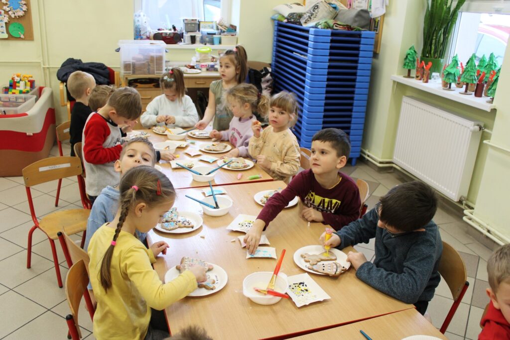 Dzieci z przedszkola w Stryszowie siedzą lub stoją przy stoliku i układają wzorki na pierniczkach przy pomocy ozdób rozłożonych na stoliku na papierowych tackach.