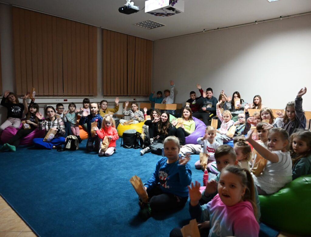 trzydzieścioro dzieci, które siedzą na dywanie, pufach i krzesłach podczas Wieczoru Filmowego z GOK.