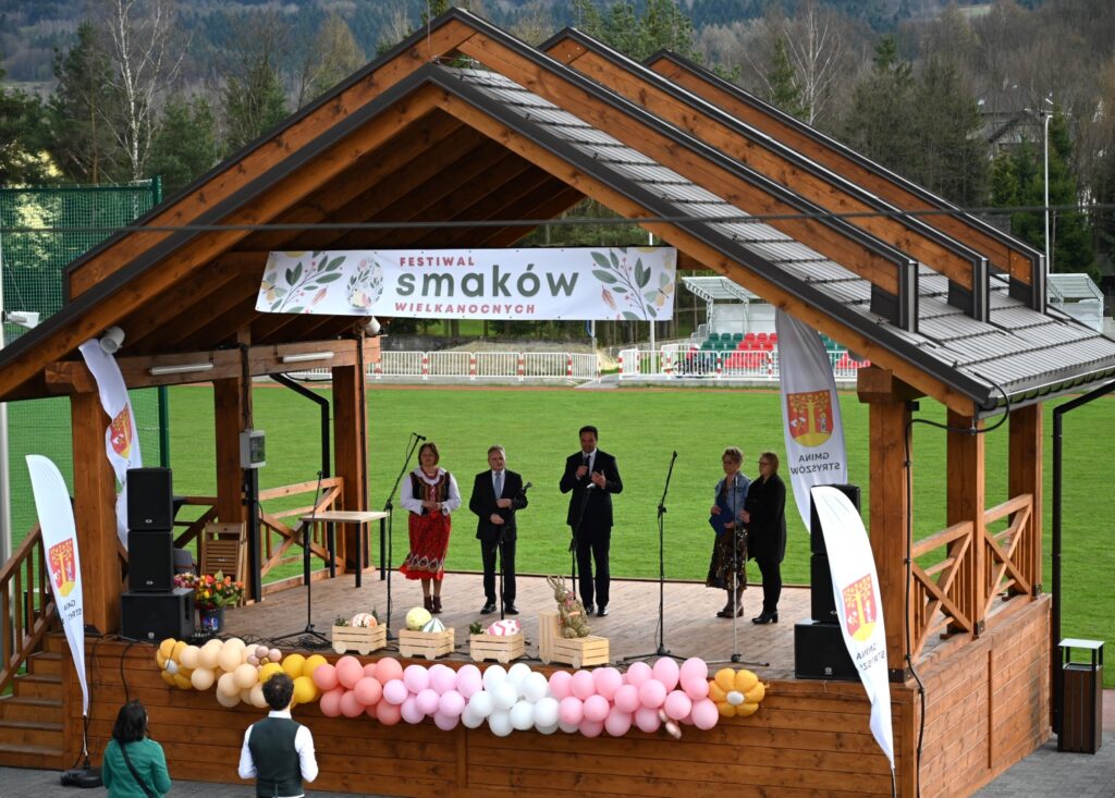 5 osób stojących na drewnianej scenie i witających przybyłych gości Festiwalu Smaków Wielkanocnych.